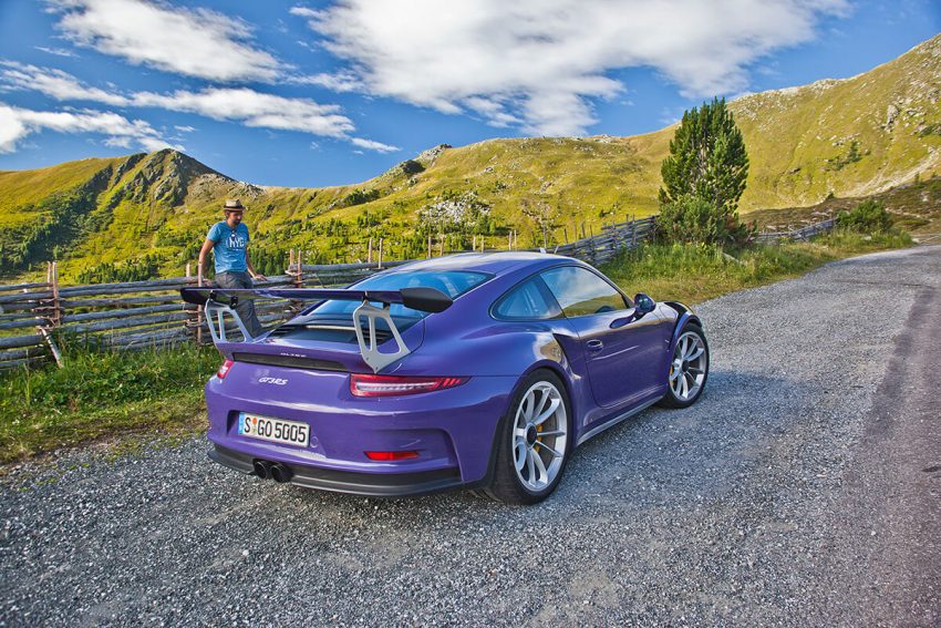 Ein lila Porsche 911 auf einer Bergstraße. Am Straßenrand steht ein Mann mit Hut. © AUTOaktuell