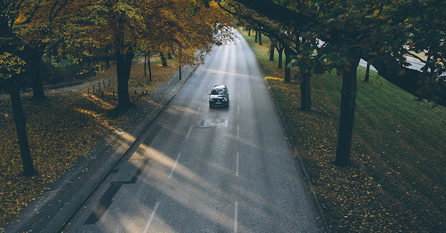 Ein Auto auf einer Straße im Herbst. Am Rand gibt es Laubbäume und auf der Straße liegt teilweise Laub. 