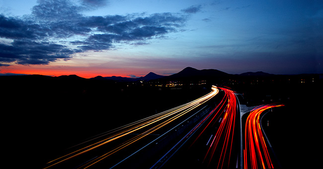 Autobahn in der Nacht. Die Scheinwerfer der Fahrzeuge sind gut sichtbar. Im Hintergrund ein Sonnenauf- oder -untergang. © pexels.com